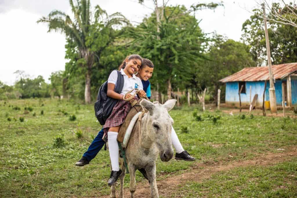 Un frère et une soeur montent sur un âne. La jeune fille porte un uniforme scolaire et un sac à dos.