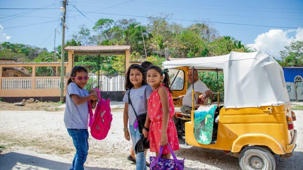 Trois filles, des amies, s'apprêtent à monter à bord d'un véhicule jaune tuk-tuk, qu'un homme conduit.