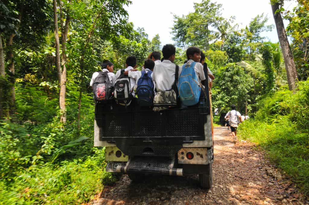 Les écoliers portant des sacs à dos roulent à l'arrière d'un camion sur un chemin de terre dans une zone rurale