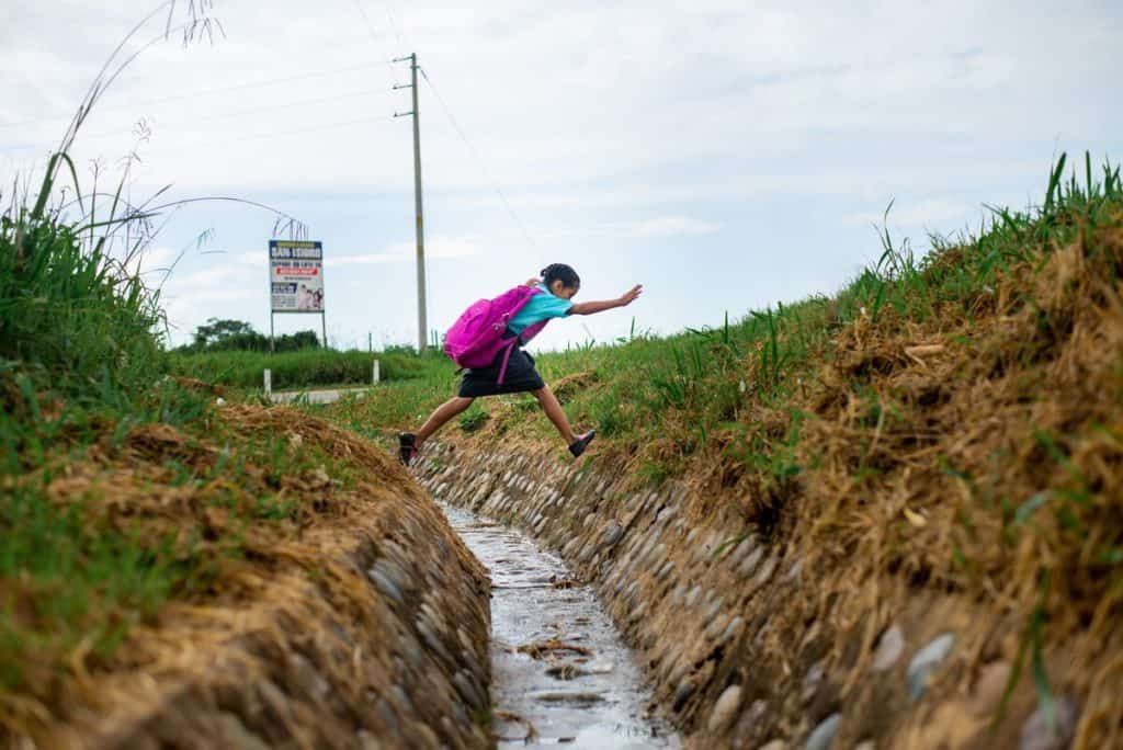 Une fille portant une chemise bleue et un sac à dos rose saute à travers un fossé. Il y a de l'eau qui coule dans le fossé.