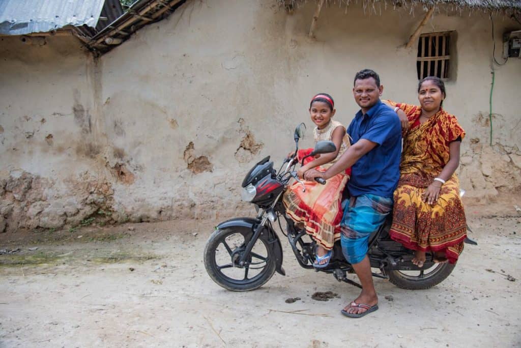 Une jeune fille est assise à l'avant d'une moto. Derrière elle se trouvent son père et sa mère. Ils portent des vêtements colorés.