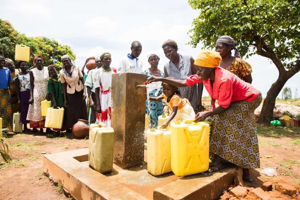 Une femme portant un foulard jaune et une chemise rouge avec une jupe longue à motifs sourit alors qu'elle va chercher de l'eau propre à un robinet extérieur extérieur de la communauté dans son récipient en plastique jaune D'autres femmes se tiennent en file indienne derrière elle pour également aller chercher l'eau courante propre.  Une fille vêtue d'une robe orange et blanche aide sa mère à collecter l'eau qui sauve des vies.