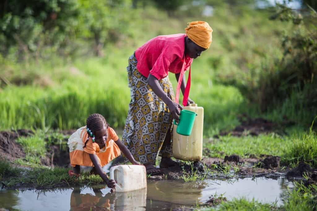 Femme portant un foulard jaune sur la tête, une chemise rouge et une jupe longue à motif dans une grande flaque d'eau où l'eau de pluie s'est accumulée sur le sol.