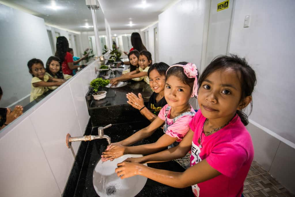 Plusieurs filles sont vues ici dans la salle de bain du centre étudiant.  Ils sourient et regardent la caméra en se lavant les mains.