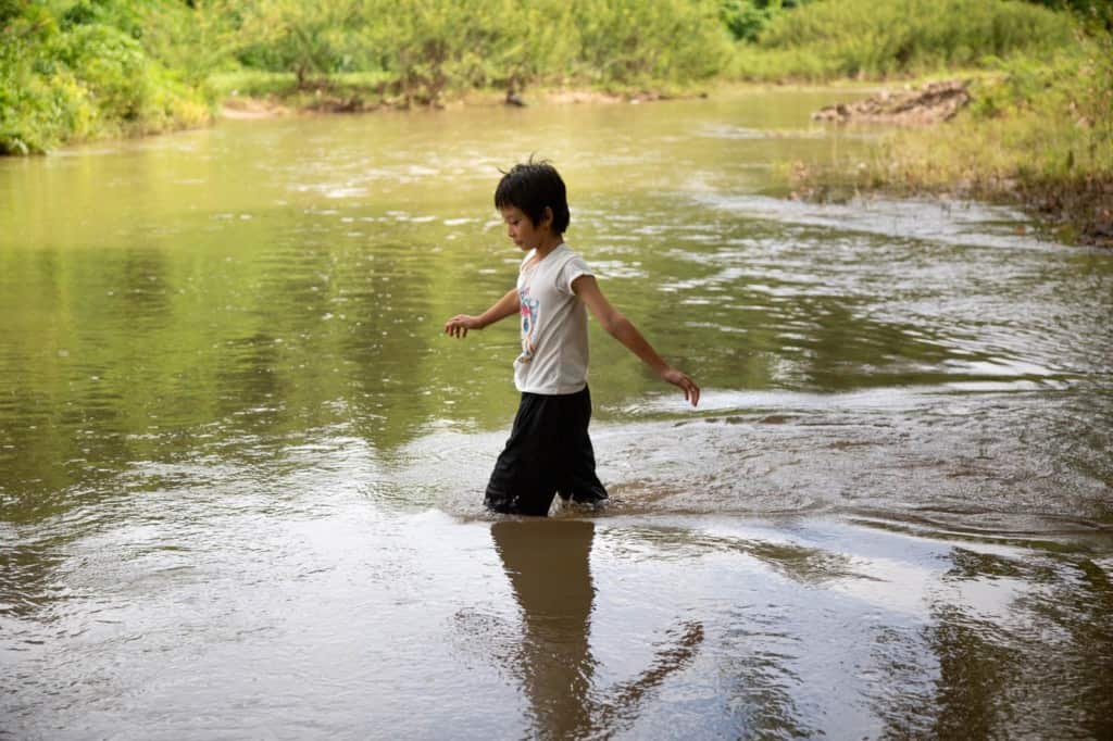 Fille vêtue d'une chemise blanche et d'un short noir.  Elle entre dans un ruisseau près de son village.