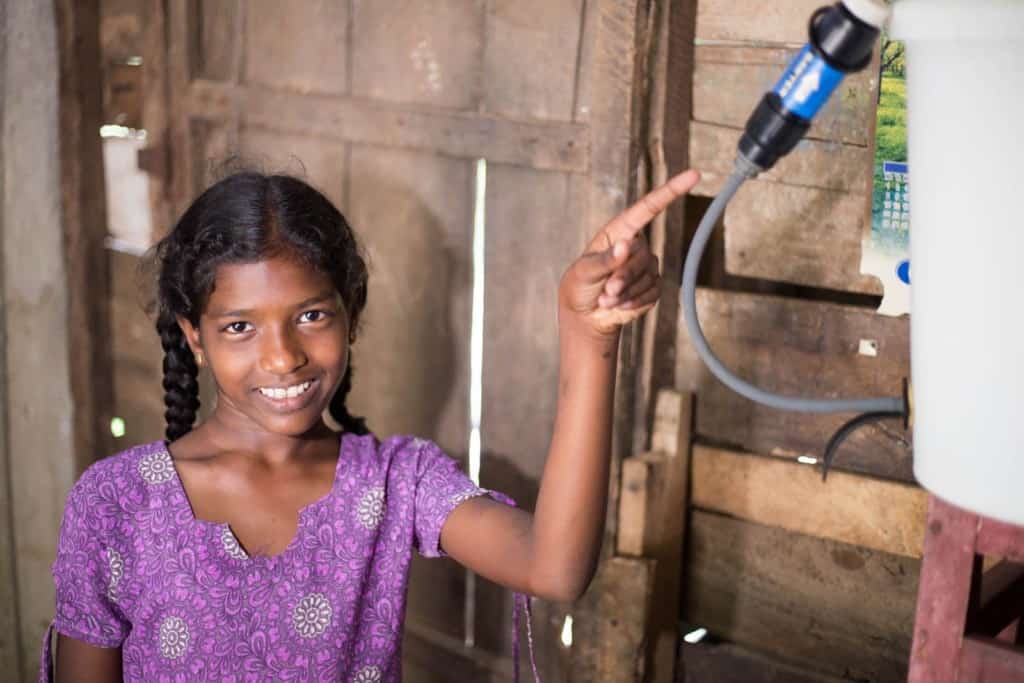 Fille debout et pointant vers le filtre Sawyer.  Elle porte un haut violet et sourit à la caméra.