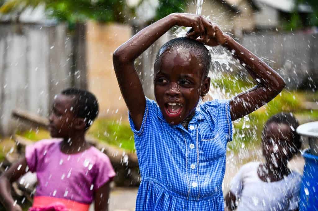 Fille vêtue d'une robe à carreaux bleu et blanc.  Elle joue dans l'eau qui va du tuyau au forage du centre Compassion.  L'eau coule sur sa tête.  D'autres enfants sont à l'arrière-plan.