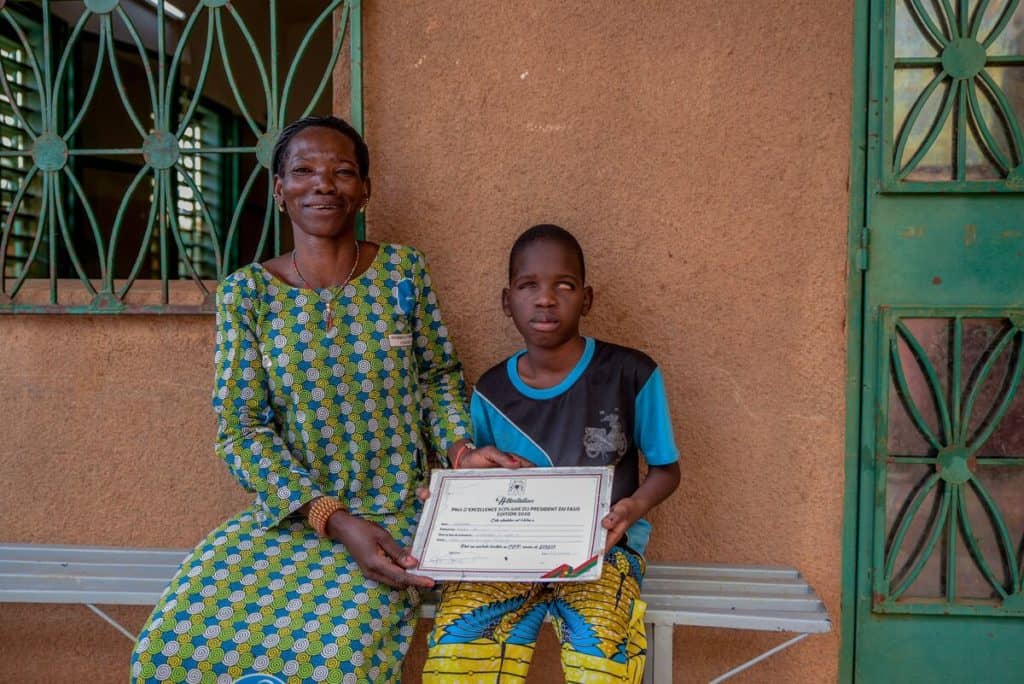 Une mère forte vêtue d'une robe verte, bleue et blanche sourit fièrement alors qu'elle et son fils brandissent un certificat de reconnaissance d'excellence académique du président du Burkina Faso.