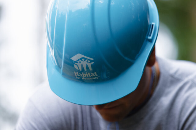 Une personne portant un casque bleu d'Habitat pour l'humanité, la tête baissée, le visage obstrué par le chapeau qui comporte un logo HFH blanc